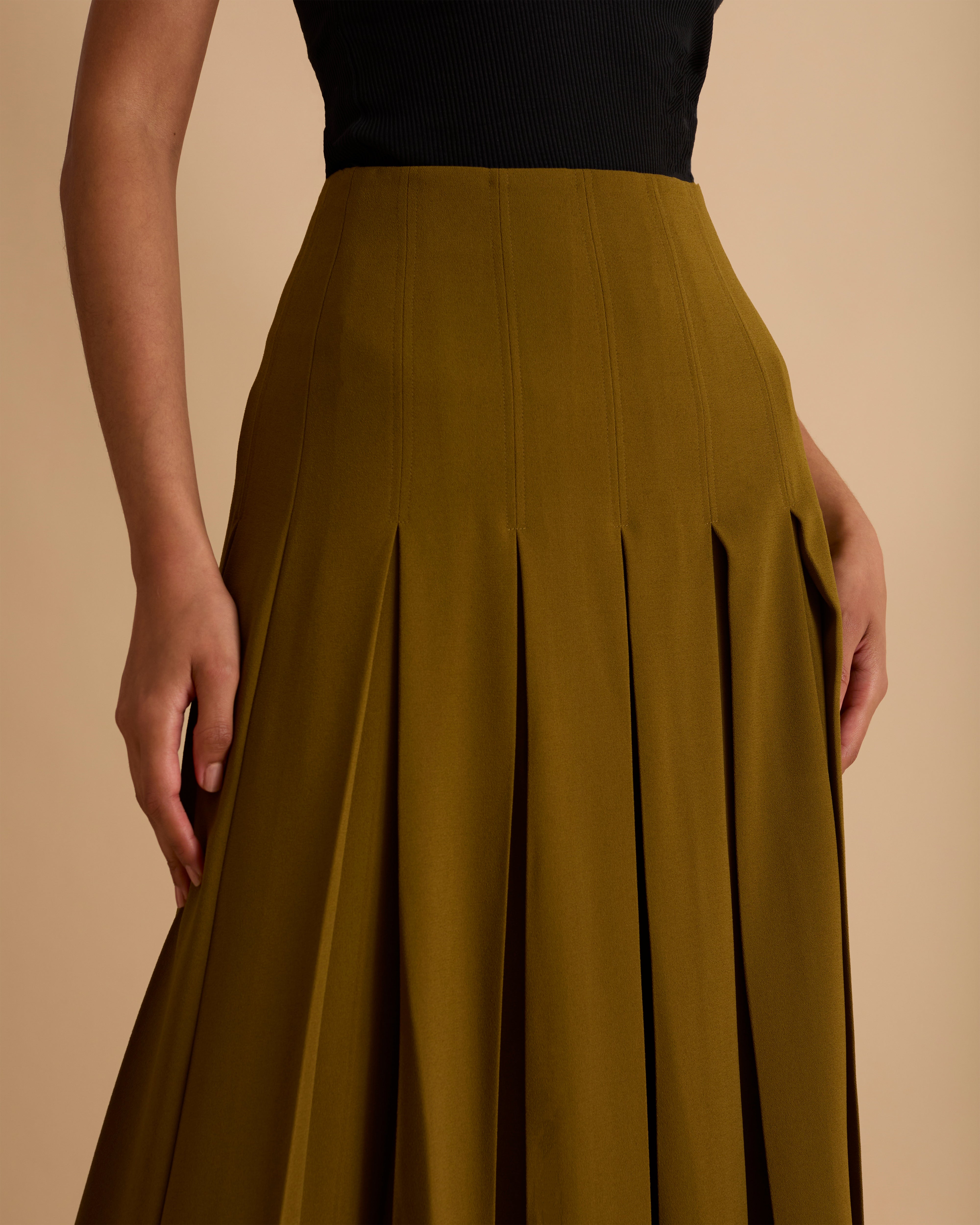 Model wearing Florere deep pleated midi skirt in olive green, wearing black boots and matching sleeveless waistcoat with gold chunky necklace on a beige neutral backdrop