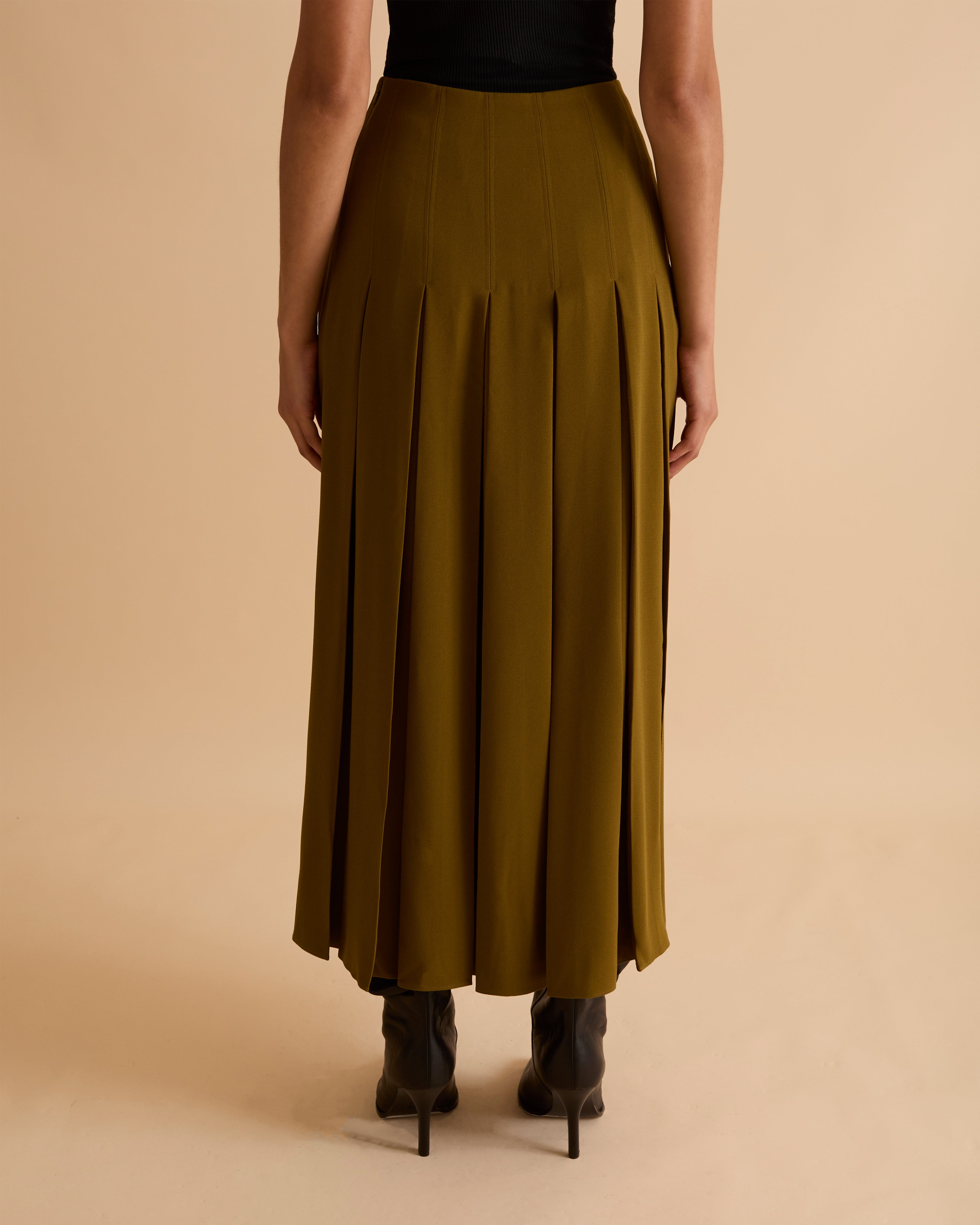 Model wearing Florere deep pleated midi skirt in olive green, wearing black boots and matching sleeveless waistcoat with gold chunky necklace on a beige neutral backdrop