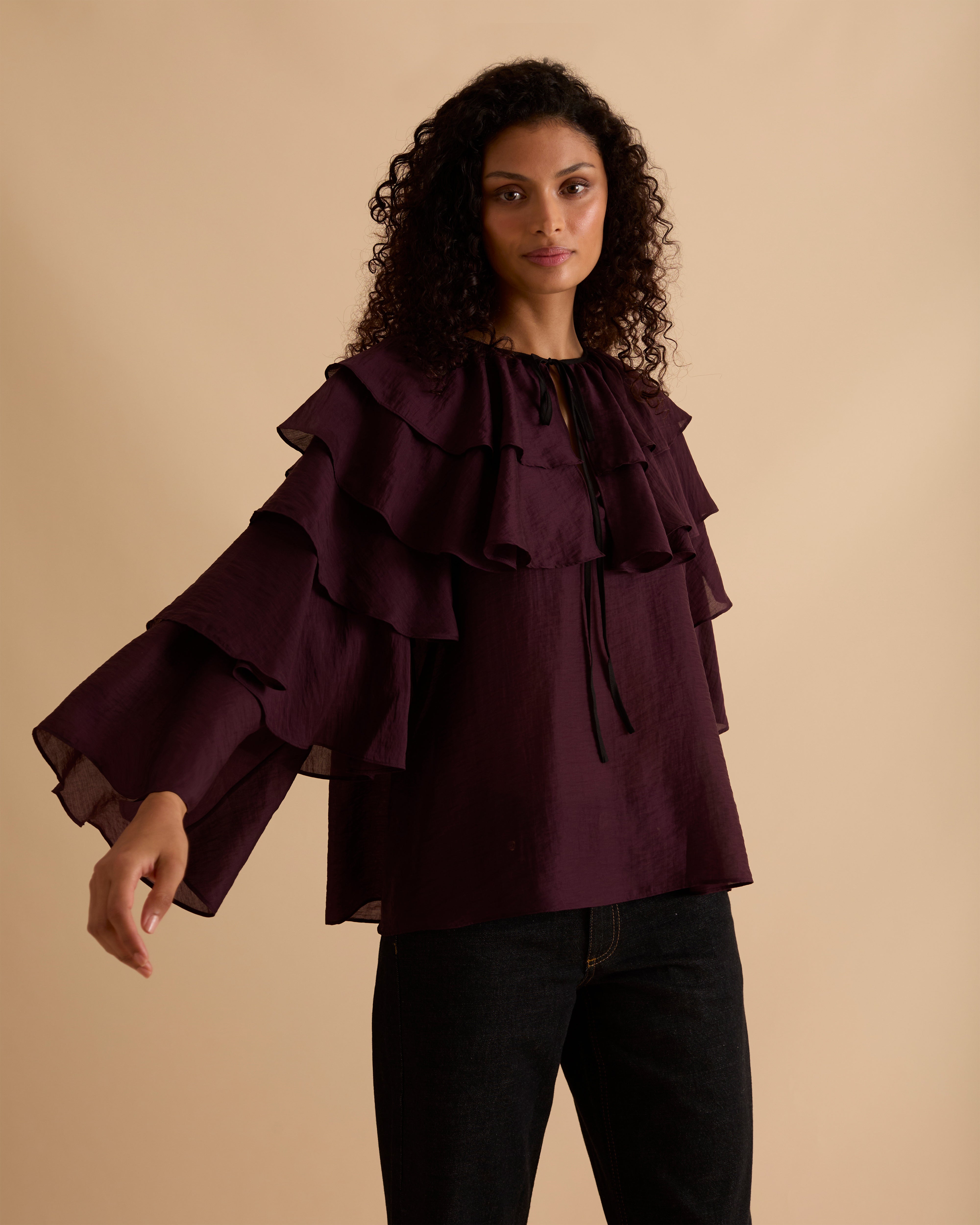 Model wearing Florere multi tiered and ruffle burgundy top in billowing lightweight fabric, loosely fitted with long sleeves and ties at neck. Wearing with black jeans and stilettos against a beige neutral backdrop