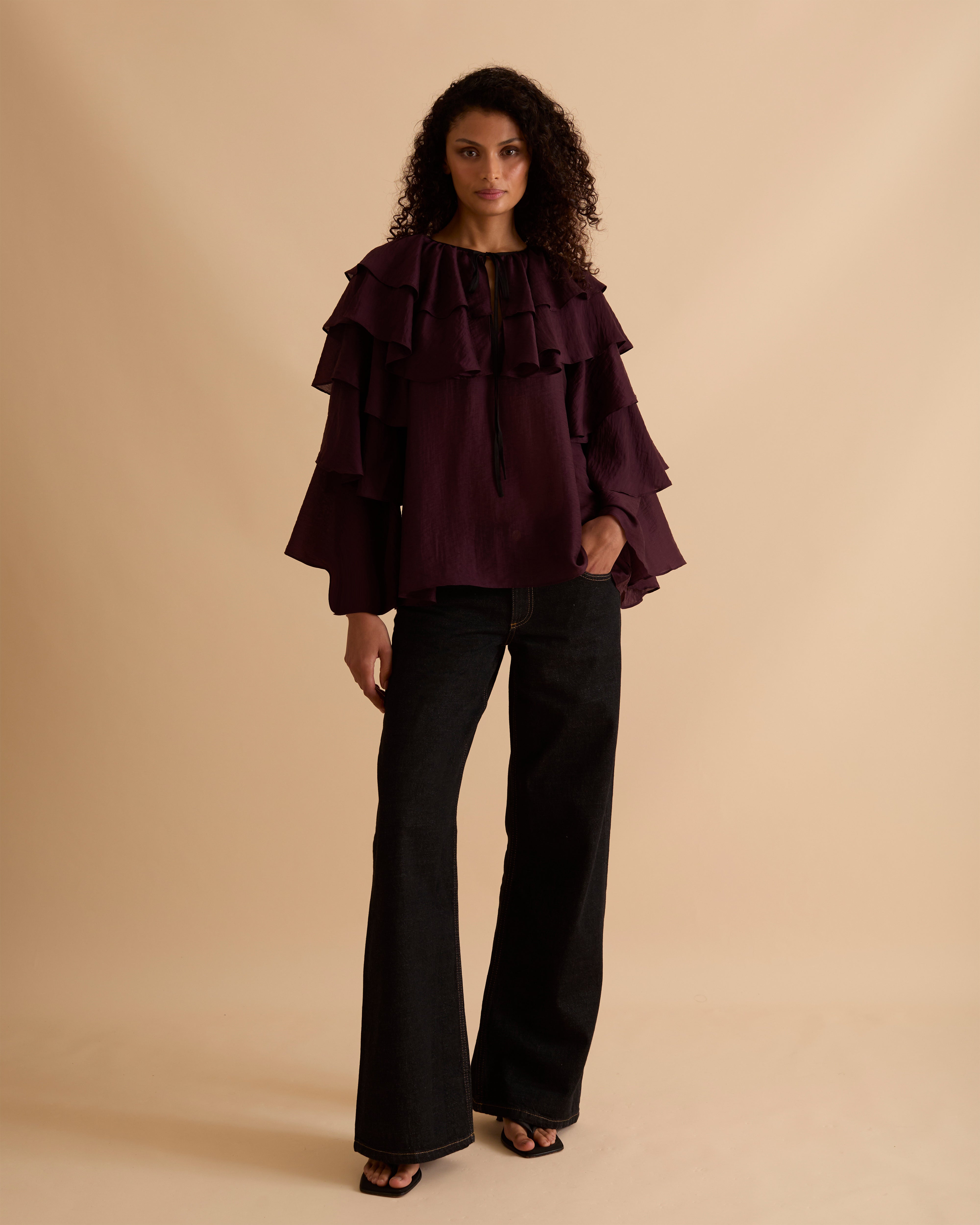 Model wearing Florere multi tiered and ruffle burgundy top in billowing lightweight fabric, loosely fitted with long sleeves and ties at neck. Wearing with black jeans and stilettos against a beige neutral backdrop