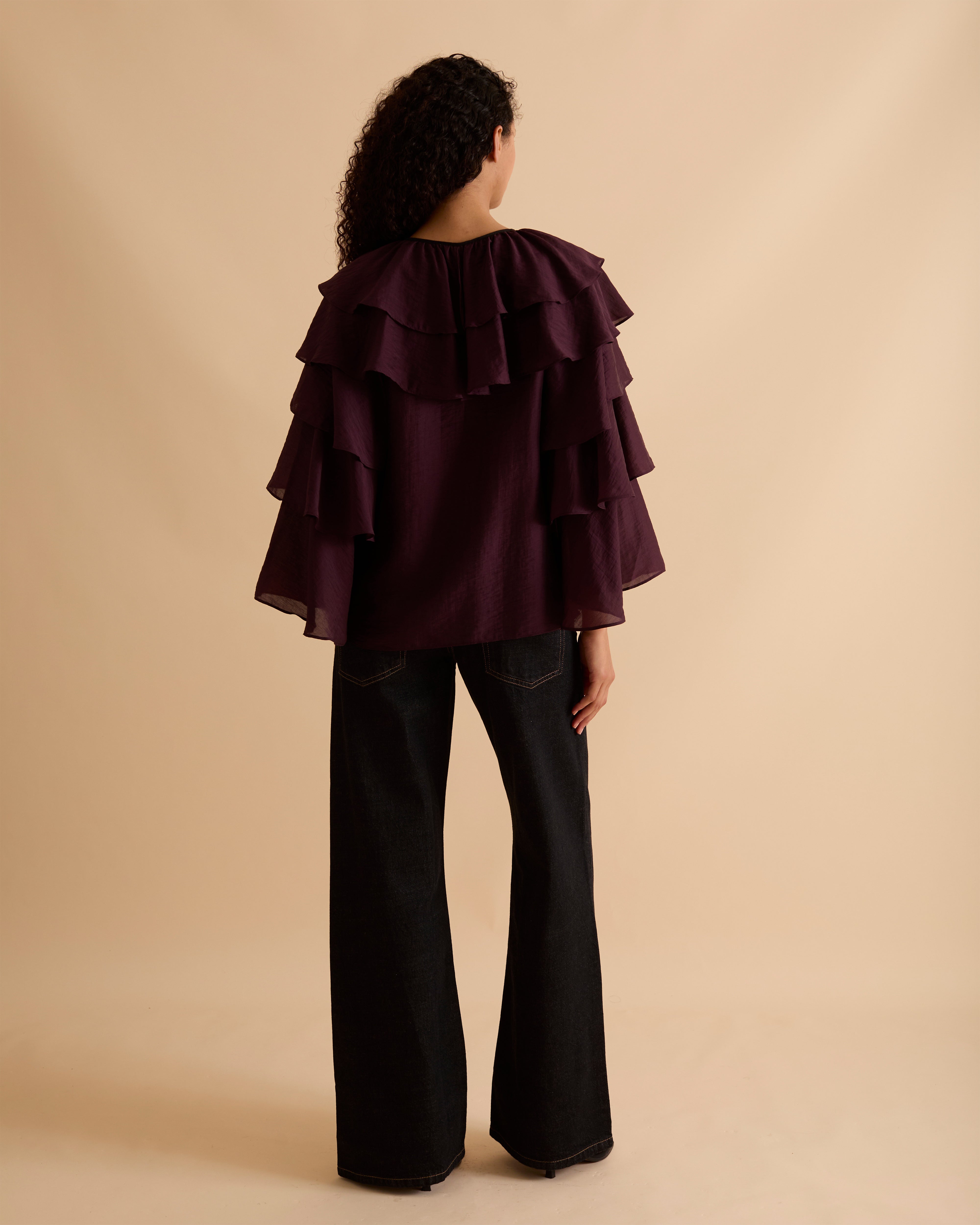 Model wearing Florere multi tiered and ruffle burgundy top in billowing lightweight fabric, loosely fitted with long sleeves and ties at neck. Wearing with black jeans and stilettos against a beige neutral backdrop