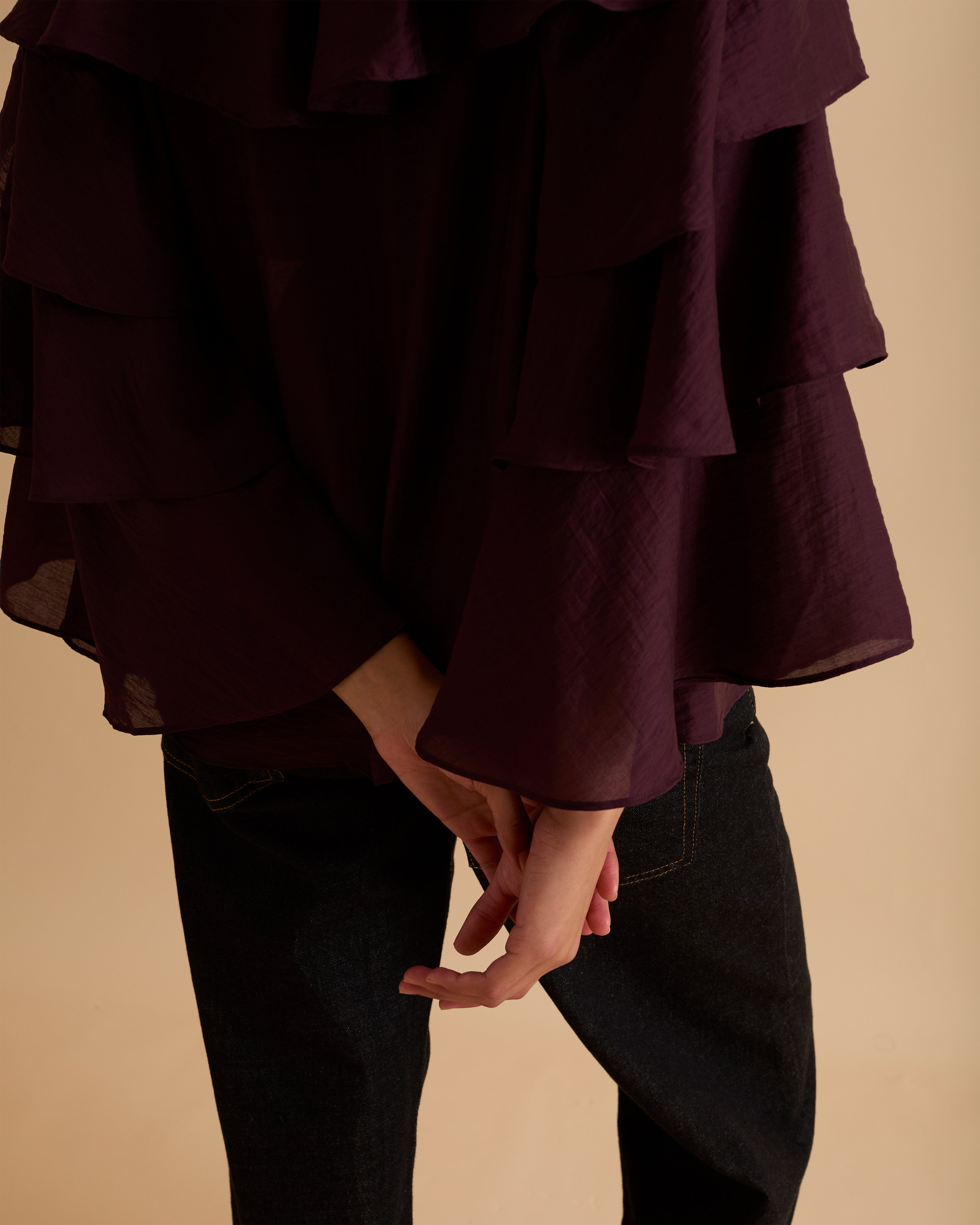Model wearing Florere multi tiered and ruffle burgundy top in billowing lightweight fabric, loosely fitted with long sleeves and ties at neck. Wearing with black jeans and stilettos against a beige neutral backdrop