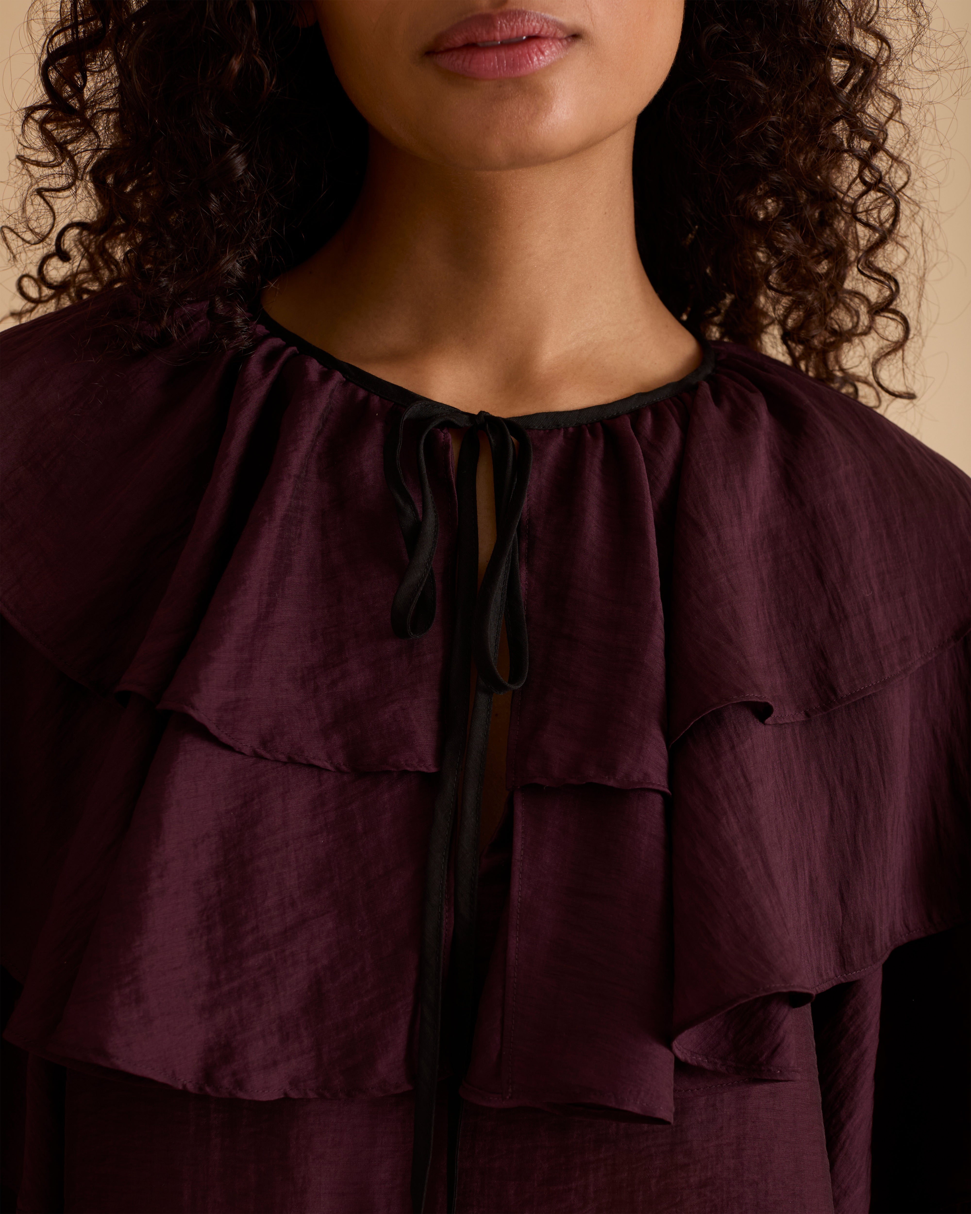 Model wearing Florere multi tiered and ruffle burgundy top in billowing lightweight fabric, loosely fitted with long sleeves and ties at neck. Wearing with black jeans and stilettos against a beige neutral backdrop