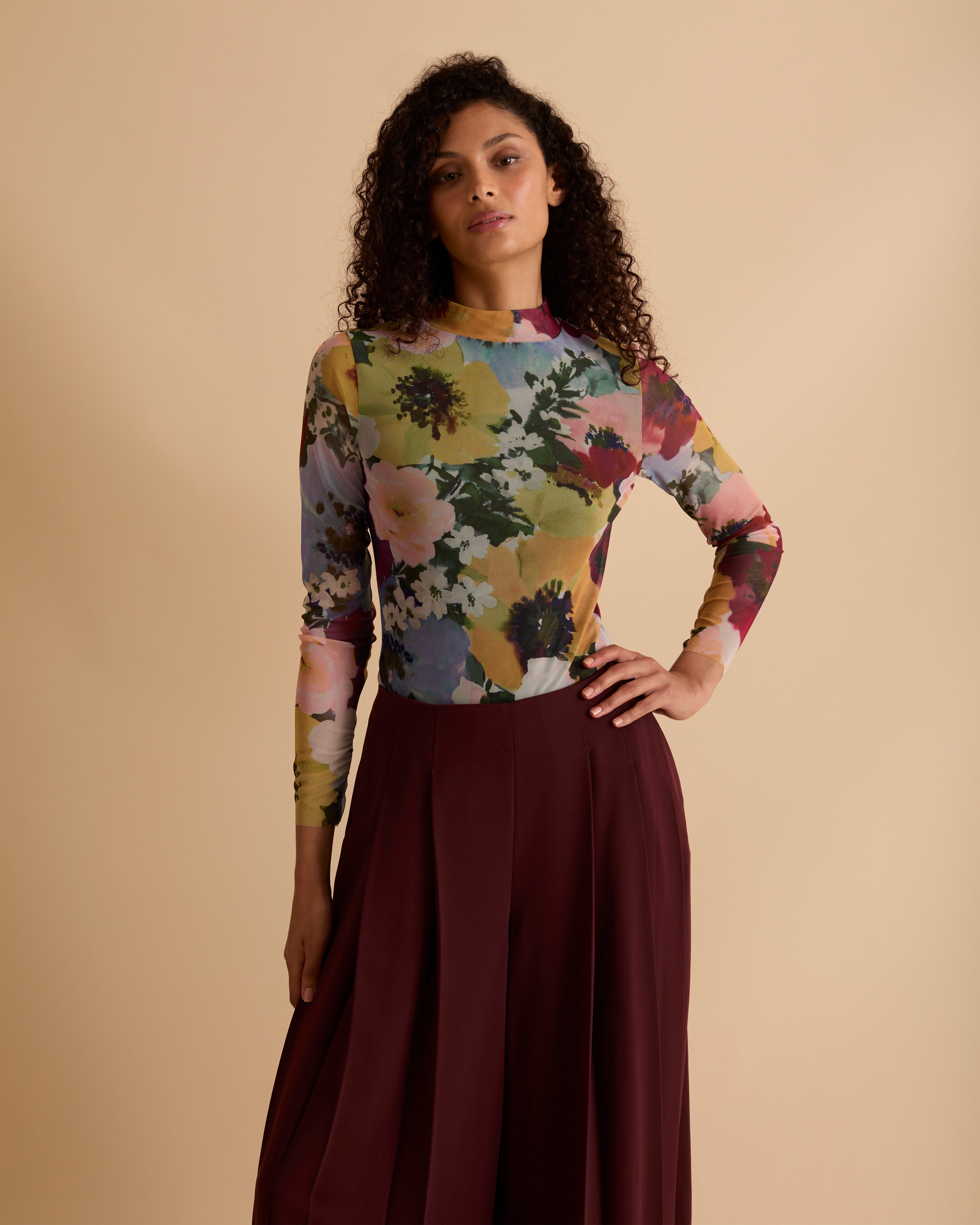 Model wearing Florere floral print mesh top with high neck and long fitted sleeves. Wearing with Florere wide leg pleated burgundy trousers against a neutral back drop