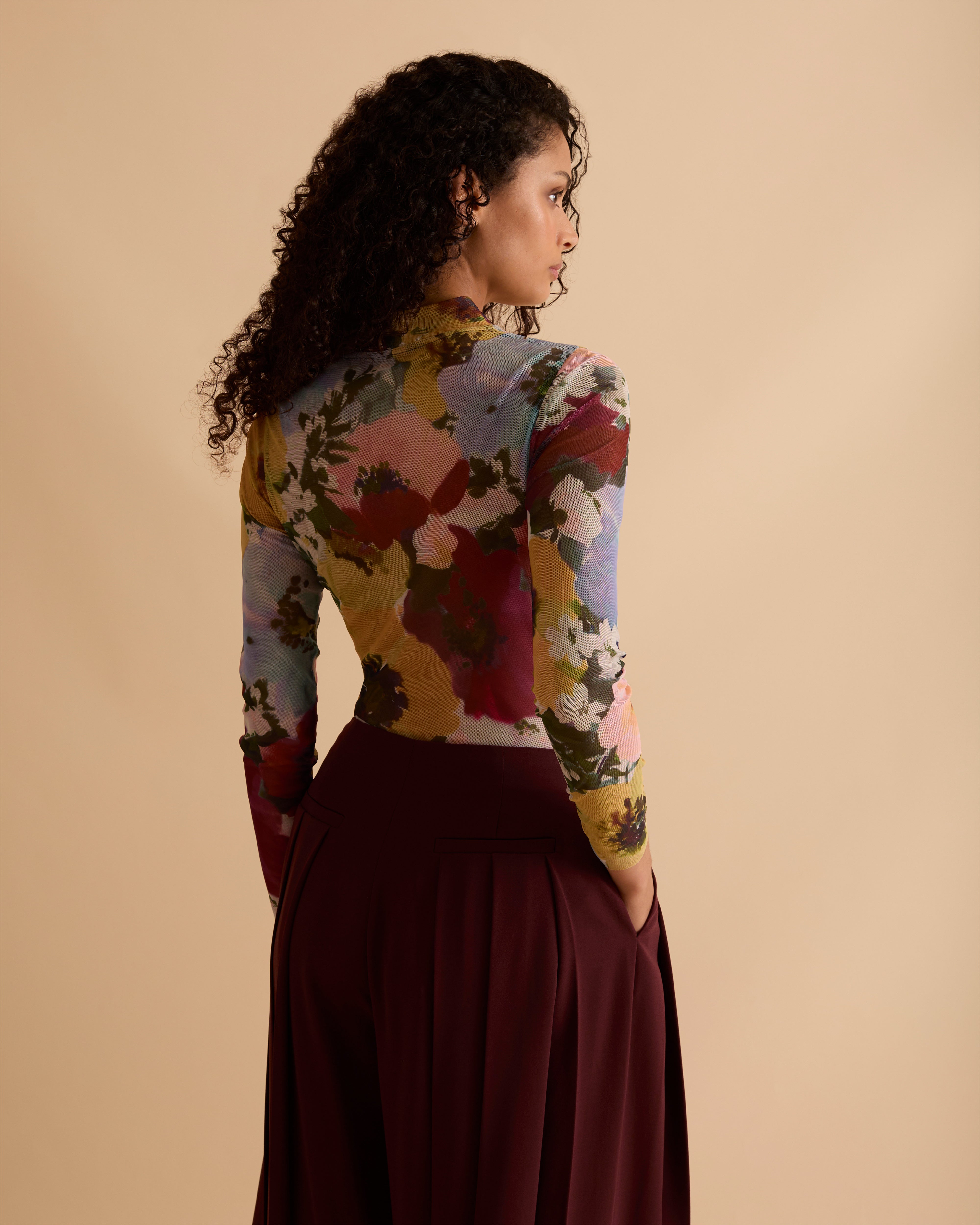 Model wearing Florere floral print mesh top with high neck and long fitted sleeves. Wearing with Florere wide leg pleated burgundy trousers against a neutral back drop